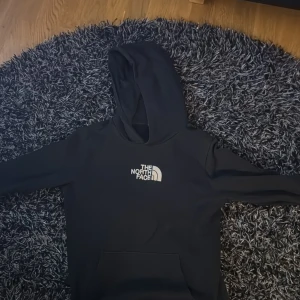 Svart hoodie från The North Face - Säljer en svart hoodie från The North Face med vit logga på bröstet. Den har en klassisk design med huva och en stor ficka framtill. Perfekt för en avslappnad stil.