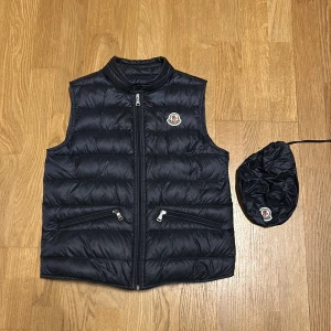 Svart Monclerväst för barn - Moncler gui väst för barn. Bra skick, använd några gånger inte sliten. Finns dock en liten slitning ovanför höger ficka därav det billiga priset. Storlek 12/152cm (Bara att höra av sig vid frågor kring västen)