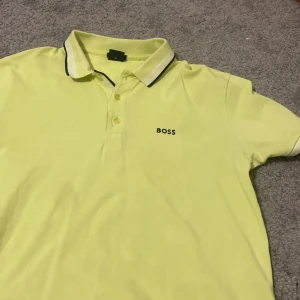 Gul pikétröja från Boss - Snygg gul pikétröja från Boss med klassisk krage och knappar framtill. Tröjan har en broderad logotyp på bröstet och text på kragen. Perfekt för en stilren look.storlek m