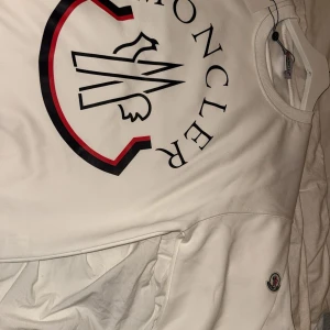 Vit sweatshirt från Moncler - Säljer en stilren vit sweatshirt från Moncler med stort tryck på framsidan i svart och rött. Tröjan har långa ärmar och en rund halsringning. Perfekt för en casual look med en touch av lyx.