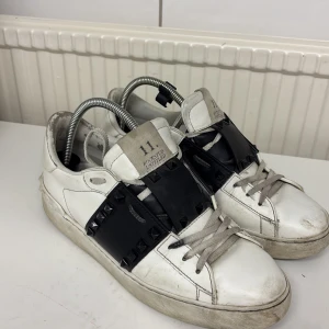 Vita sneakers med svarta detaljer - Snygga vita sneakers från Valentino med svarta detaljer och nitar. Skorna har snörning och en unik design med breda svarta band över ovansidan. Perfekta för en trendig look. Skick 8/10 saknar inte några nitar