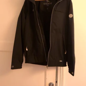 Svart jacka från Snoot - Snygg svart jacka från Snoot med dragkedja och långa ärmar. Jackan har en stilren design med diskreta detaljer och en knapp vid ärmsluten. Perfekt för en stilren look. Storlek M