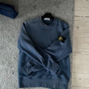 Stone island tröja  - Säljer en snygg blå sweatshirt från Stone Island med deras ikoniska märke på ärmen. Tröjan har en klassisk rund hals och långa ärmar. Perfekt för en stilren och avslappnad look. Storlek M men passar S också. Köpt från Plick. Tror att den är äkta för scanen funkar. Säljer billigt för är inte säker om den är äkta men tror de