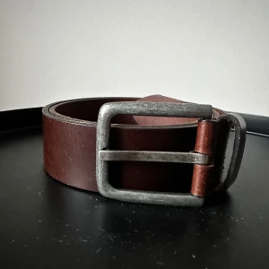 Brunt skärp med metallspänne  - Säljer ett stilrent brunt skärp i skinn med ett robust metallspänne. Perfekt för att ge en klassisk touch till din outfit. Skärpet har flera hål för justerbar passform. Sparsamt använt. Längd 90 cm.