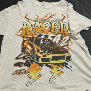 Vit t-shirt med racertryck - Cool vit t-shirt med ett stort tryck av en racerbil och texten 'Kansas City Racer 42' i orange och gul. Perfekt för bilentusiaster som vill sticka ut med en unik design. T-shirten har korta ärmar och en avslappnad passform.