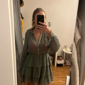 Grön klänning med volanger - Snygg grön klänning med volanger och är  v-ringad. Klänningen har långa ärmar med knappar framtill. Endast använd en gång. Skriv för fler bilder💕