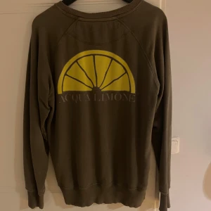 Olivgrön tröja från Acqua Limone - Säljer en olivgrön tröja från Acqua Limone med tryck av en gul citronhalva på baksidan och logotyp på framsidan. Tröjan har långa ärmar och ribbade muddar vid ärmslut och nederkant. Perfekt för en avslappnad stil. STRL M