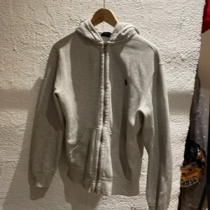 Grå hoodie från Ralph Lauren - Säljer en stilren grå hoodie från Ralph Lauren med dragkedja och det klassiska logotypbroderiet på bröstet. Perfekt för en avslappnad look. Hoodien har långa ärmar och en bekväm passform. Har lite fläckar men syns typ inte om man kolar från håll 