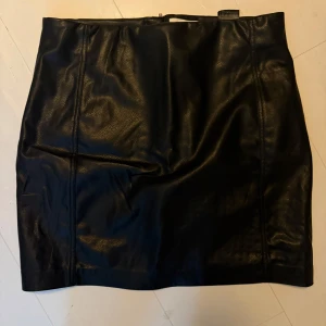 Svart skinnkjol - Snygg svart skinnkjol med en stilren design. Kjolen har en tight passform och är perfekt för att ge en edgy look. Den är kort och har en dragkedja baktill för enkel på- och avtagning.