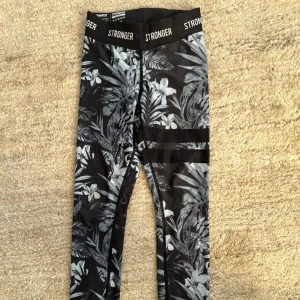 Svarta leggings med blommönster från Stronger - Snygga svarta leggings från Stronger med ett stilrent blommönster i grått och vitt. De har en hög midja och är tillverkade i en mjuk och stretchig polyesterblandning. Mönstret heter Pompeii. Perfekta för träning eller en avslappnad dag.