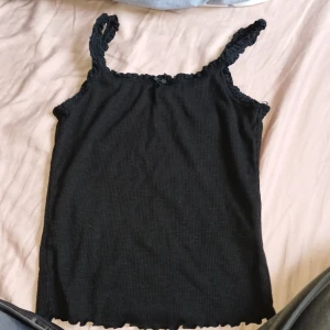 Svart ribbad linnetopp från H&M Divided - Säljer en svart ribbad linnetopp från H&M Divided. Toppen har smala axelband och volanger vid kanterna. 