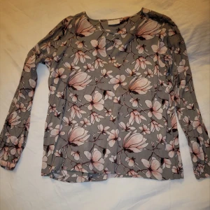 Grå blommig blus - Säljer en elegant grå blus med vackert blommönster i rosa och vitt. Blusen har långa ärmar och en rund halsringning med ett litet sprund, perfekt för en stilren look.
