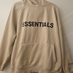 Beige hoodie från Essentials - Beige hoodie från Essentials med svart text på bröstet. Den har en klassisk känguruficka och ribbade muddar vid ärmslut och nederkant.