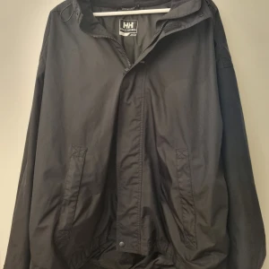 Svart vindjacka från Helly Hansen - Svart vindjacka från Helly Hansen med huva som kan vikas in.