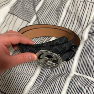 Svart bälte med silverfärgat spänne - Snyggt svart bälte med ett stort, runt silverfärgat spänne. Bältet har ett diskret mönster och är perfekt för att ge en stilren touch till din outfit. Passar bra till både jeans och kostymbyxor.