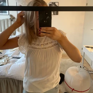 Vit blus med spetsdetaljer - Säljer en söt vit blus med spetsdetaljer på ärmarna och runt halsen. Blusen har korta ärmar och en liten knappöppning i ryggen. Perfekt för en somrig look! Använd fåtal gånger, inga defekter
