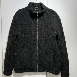 Svart teddyjacka - HM - Mysig svart teddyjacka med dragkedja framtill. Perfekt för kyliga dagar med sin fluffiga och varma design. Jackan har långa ärmar och två praktiska fickor på sidorna. Från HM.