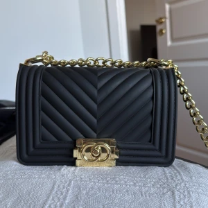 Svart quiltad axelväska med guldkedja - Snygg svart axelväska med quiltat mönster och en elegant guldkedja. Väskan har en stilren design med ett dekorativt guldfärgat spänne framtill. Perfekt för att ge din outfit en lyxig touch.