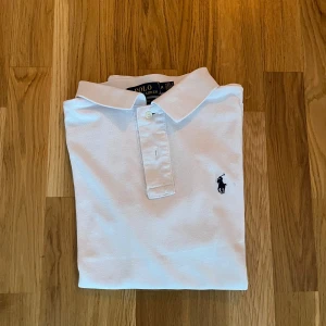 Vit pikétröja från Ralph Lauren - Säljer en klassisk vit pikétröja från Ralph Lauren med den ikoniska loggan broderad på bröstet. Tröjan har en stilren design med krage och knappar framtill. Perfekt för en avslappnad men ändå stilfull look. Pris kan diskuteras