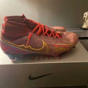  fotbollsskor från Nike - Snygga lila fotbollsskor från Nike med gula och blå detaljer. Skorna har en hög ankelkrage och snörning för optimal passform. Perfekta för fotbollsplanen med sin moderna design och funktionalitet.