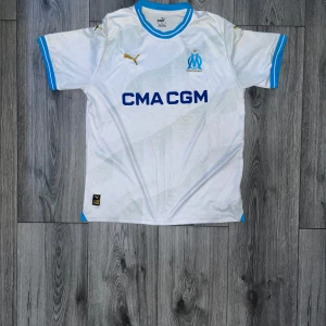 Marseille tröja  - Säljer en vit fotbollströja från Puma med blå detaljer och korta ärmar. Tröjan har klubbmärket på bröstet och sponsorloggan 'CMA CGM' tryckt på framsidan. Detaljer i guld och texten 'PETRA GENESIS MASSALIA' på baksidan.
