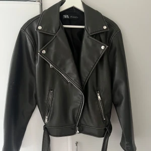 Svart skinnjacka från Zara - Snygg svart skinnjacka från Zara med dragkedja och knappar. Jackan har en klassisk bikerstil med bälte i nederkant och långa ärmar. 