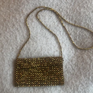 Guldig axelväska - Snygg guldig axelväska med glittrande paljetter. Perfekt för att lysa upp din outfit. Väskan har en elegant kedjerem som ger en extra touch av glamour.