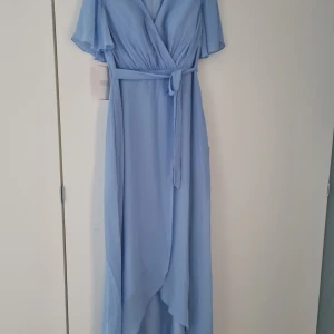 Ljusblå klänning - Helt ny Elegant ljusblå omlottklänning med v-ringning och kortärmade ärmar. Klänningen har ett knytband i midjan och en asymmetrisk nederdel som ger en fin rörelse. Perfekt för sommarens festligheter, bröllop eller bal.