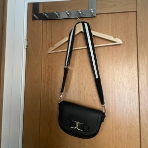 Svart axelväska med gulddetalj - Snygg svart axelväska från even & odd med en elegant gulddetalj på framsidan. Väskan har en justerbar axelrem med svart och vit rand. Perfekt för att ge en stilren touch till din outfit.