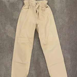 Beige byxor från Bershka - Snygga beige byxor från Bershka i straight modell. De har en högmidjad design med knappar och dragkedja framtill. Använda endast två gånger.