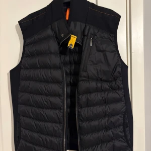 Svart dunväst, parajumpers - Snygg svart dunväst från Parajumpers med dragkedja och en praktisk bröstficka. Västen har en quiltad design och en hög krage med en gul detalj. Perfekt för lager-på-lager under kyliga dagar. Den är typ aldrig använd därför säljer jag den. Den är i storlek Xl och den är äkta men fick den i present så har tyvärr inte kvitto tveka inte på att höra av er vid frågor 