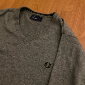 Grå tröja från Fred Perry - Säljer en stilren grå tröja från Fred Perry med v-ringning och broderad logga på bröstet. Perfekt för en avslappnad men snygg look. Storlek S. Skick 10/10. Använd endast ett fåtal gånger. Originalpris 59 dollar.