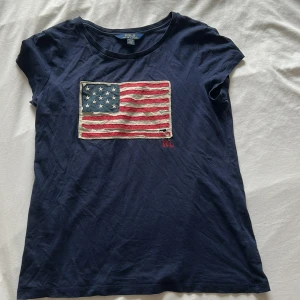 Mörkblå t-shirt från Ralph Lauren - Snygg mörkblå t-shirt från Ralph Lauren med ett broderat amerikanskt flaggmotiv på framsidan. T-shirten har korta ärmar och en rund halsringning. Perfekt för en avslappnad stil.