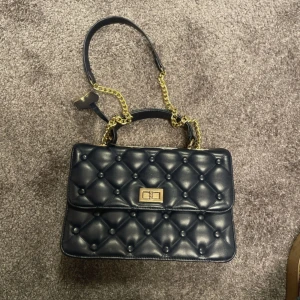 Svart quiltad axelväska från Shuberry - Snygg svart quiltad axelväska från Shuberry med guldfärgade kedjedetaljer. Väskan har en elegant låsdetalj framtill och en röd insida med dragkedjefack för extra förvaring. Perfekt för att ge din outfit en lyxig touch.