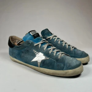 Blå Golden Goose GGDB - Snygga blå sneakers från Golden Goose. Skorna har en klassisk snörning och en kontrasterande svart häl. Perfekta för en avslappnad stil. Skick: 2,5/5. Skorna kommer med orginal boxen.