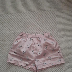 Rosa shorts med tryck - Söta rosa shorts med ett charmigt tryck av Snoopy-figurer. De har en elastisk midja för extra komfort och är perfekta för en avslappnad stil. Materialet känns mjukt och skönt mot huden.