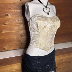 Beige korsett med broderade detaljer - Elegant beige korsett med vackra broderade mönster. Den har en strukturerad passform och en subtil glans i tyget. Perfekt för att ge en stilfull touch till din outfit.