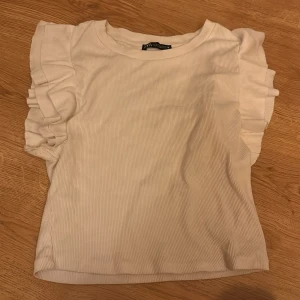 Vit ribbad topp från Zara - Säljer en söt vit ribbad topp från Zara med volangdetaljer på ärmarna. Perfekt för en stilren look. Toppen har en rund halsringning och är tillverkad i ett mjukt material.💗