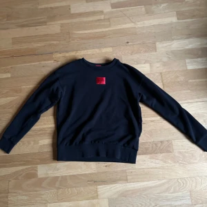 Svart tröja från Hugo - Säljer en stilren svart tröja från Hugo med en röd logga på bröstet. Tröjan har långa ärmar och en rund halsringning. Perfekt för en casual look.