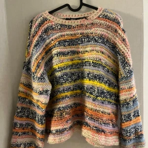 Flerfärgad stickad tröja - Säljer en mysig flerfärgad stickad tröja med ett unikt mönster i blått, gult, rosa och orange. Tröjan har långa ärmar och en avslappnad passform. Perfekt att typ att kunna dra över på sena sommarkvällar. 