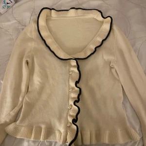 Vit kofta med volangdetaljer - Charmig vit kofta med svarta volangdetaljer längs kragen och knappslån. Koftan har långa ärmar och pärlknappar framtill. Perfekt för en elegant och stilren look. 