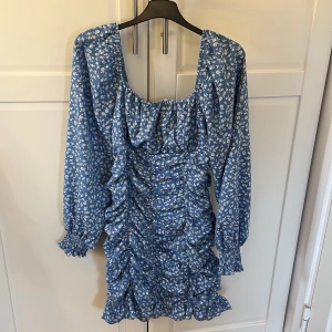 Blå blommig klänning - Charmig blå klänning med vita blommor och volanger. Den har långa ärmar och en smickrande rynkad design som ger en fin passform. Perfekt för en somrig look!