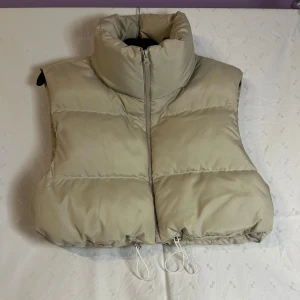 Beige dunväst från Pickk Upp - Snygg beige dunväst från Pickk Upp med dragkedja framtill. Västen har en hög krage och justerbar dragsko i nederkant för extra komfort. Perfekt för kyligare dagar. Finns några små fläckar, går att ta bort 