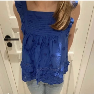 Blå ärmlös blus med volangdetaljer - Snygg blå blus med volangdetaljer och broderade blommor. Den är ärmlös och har en luftig passform, perfekt för varma dagar. Blusen har en söt volangkant längst ner som ger en extra touch.