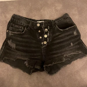 Svarta jeansshorts från Zara - Snygga svarta jeansshorts från Zara med slitna detaljer och knappar framtill. De har en avslappnad look med fransiga kanter och två bakfickor. Perfekta för en cool och avslappnad stil.