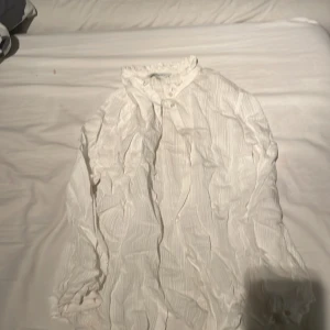 Vit blus med knappar - Säljer en stilren vit blus med knappar framtill. Blusen har långa ärmar och ett lätt rynkat tyg som ger en avslappnad look. Perfekt för en chic och enkel stil.