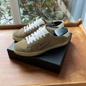 Bruna sneakers från Saint Laurent - Snygga bruna sneakers från Saint Laurent med vit snörning och vit häl. Tillverkade i mocka med märkets namn tryckt på sidan. Perfekta för en stilren look. Riktigt eftertraktade och bra pris! 8,7/10 skick!