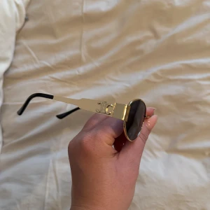 Runda solglasögon med gulddetaljer - Snygga runda solglasögon med guldfärgade bågar och mörka linser. De har en elegant design med svarta skalmar och en stilren look. Perfekta för att ge en touch av glamour till din outfit.