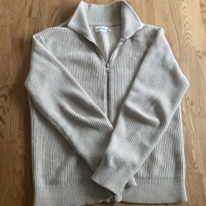 Beige ribbad tröja från East West - Säljer en stilren beige ribbad tröja från East West. Tröjan har en dragkedja framtill och en hög krage som ger en snygg look. Perfekt för kyligare dagar.