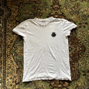Vit t-shirt från Moncler - Säljer en stilren vit t-shirt från Moncler med deras ikoniska logga på bröstet. T-shirten är tillverkad i mjuk bomull och har en klassisk passform med korta ärmar. Perfekt för en avslappnad look.